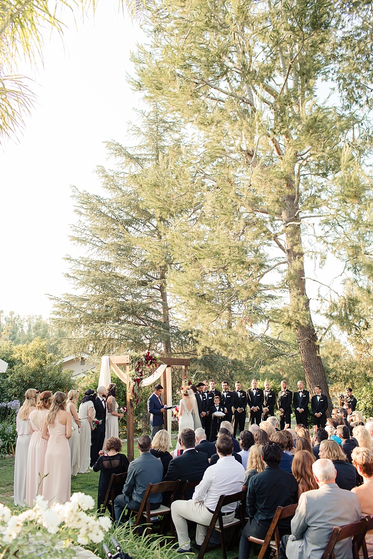 Outdoor Ceremony Bride Lace V Neck Bridal Gown Groom Bridesmaids Groomsmen Guests Wooden Decorated Arch Red Maroon Cream Roses Tall Trees Luxe Outdoor Garden Wedding in California http://figlewiczphotography.com/