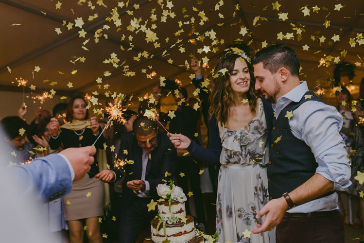 Reception Gold Confetti Sparklers Bride Patterned Floral Dress Cardigan Floral Hairpiece Groom Light Blue Shirt Dark Grey Vest Wedding Cake Guests Natural Greenery Stylish Wedding Transylvania https://raresion.com/