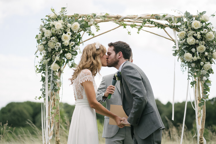Outdoor Ceremony Arch Arbour Bride Bridal Rembo Styling Lace V Neck Dress Gown Hairpiece White Cream Rose Bouquet Eucalyptus Greenery Groom Groomsman Three Piece Grey Suit Relaxed Bohemian Summer Meadow Wedding https://karibellamy.com/