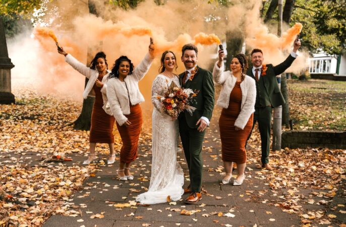 A newlywed couple and their wedding party pose happily outdoors amidst fallen autumn leaves, holding up canisters of vibrant orange smoke.