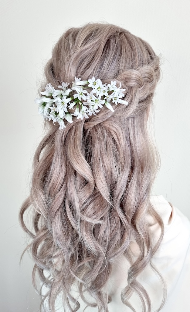 A close-up back view of a bride showcasing a half-up half-down hairstyle featuring soft waves, an intricate braid, and a cluster of fresh white wildflowers.