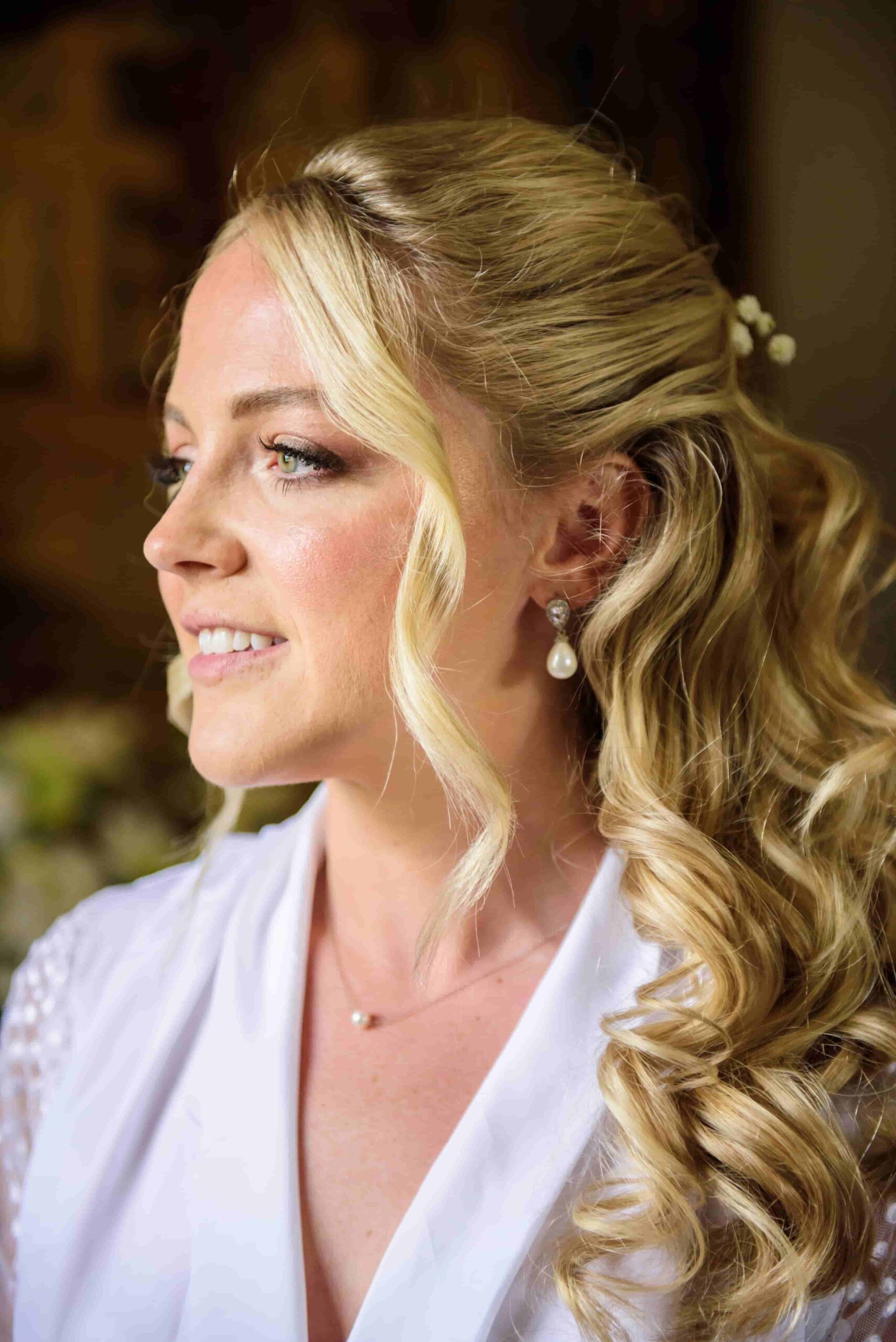 A beautiful bride getting ready on her wedding day, wearing a white robe, pearl drop earrings, and a half-up half-down hairstyle with soft waves and delicate flowers.
