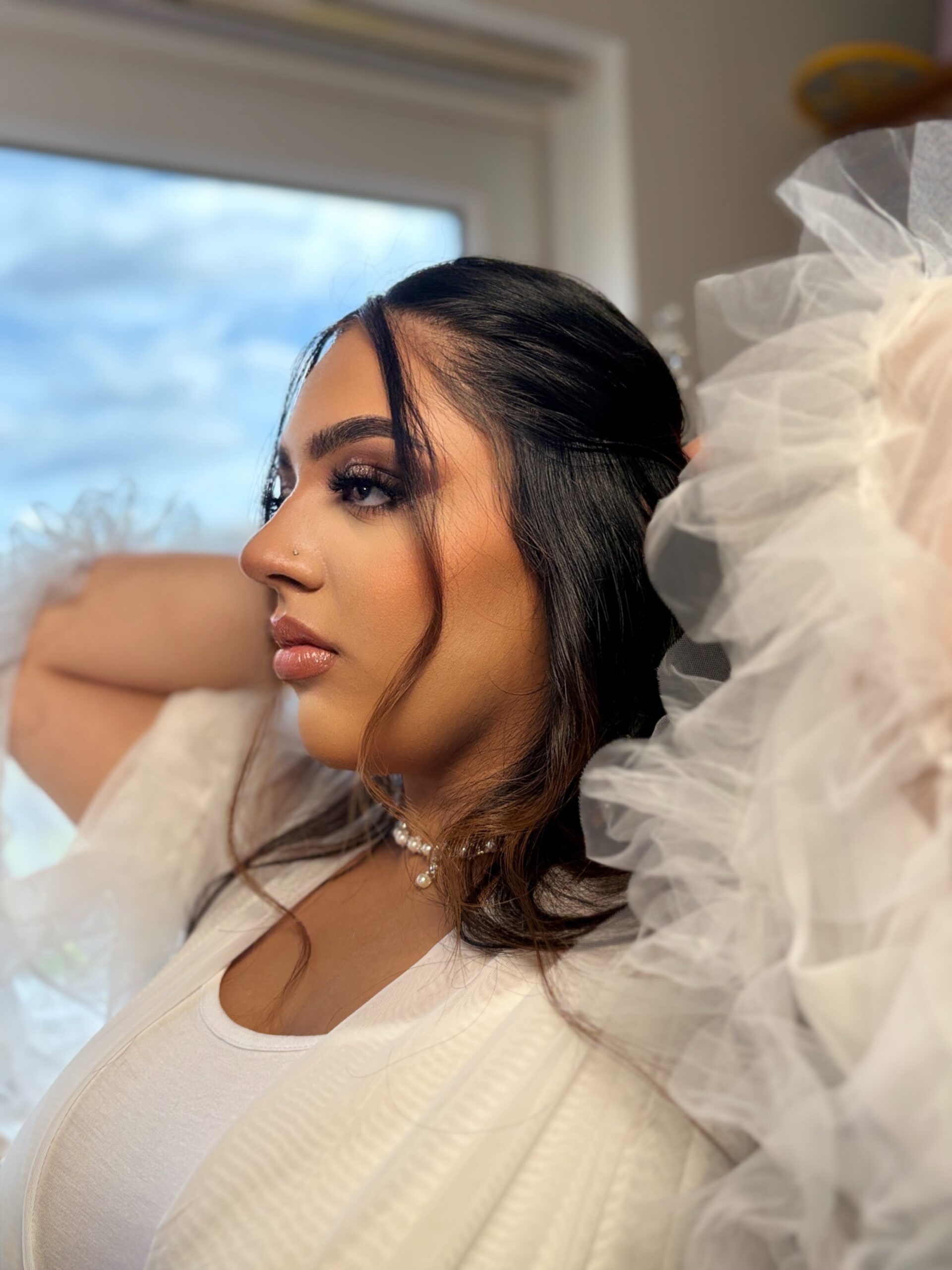 A close-up profile portrait of a bride getting ready by a window, wearing a white ruffled robe and a pearl necklace with soft glam makeup.