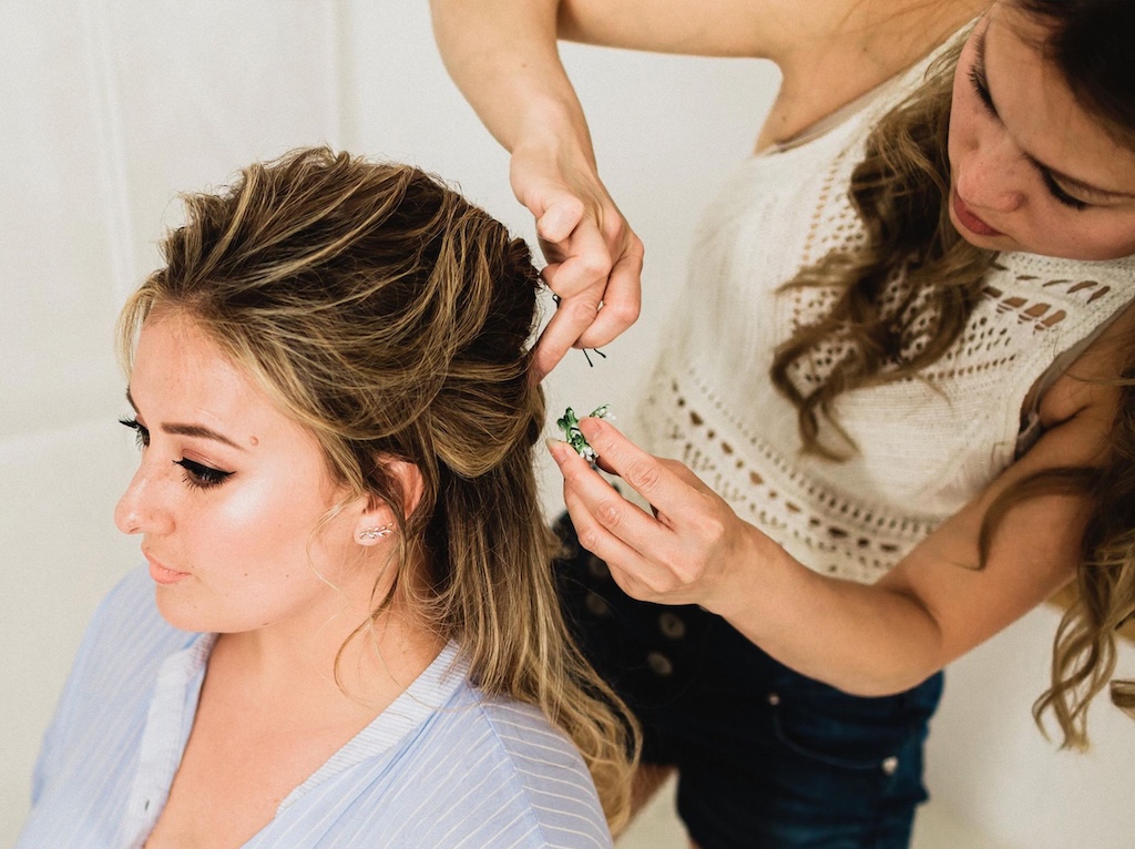 A bridal stylist carefully secures an elegant half-up hairstyle with hair pins while the bride looks on during morning preparations.