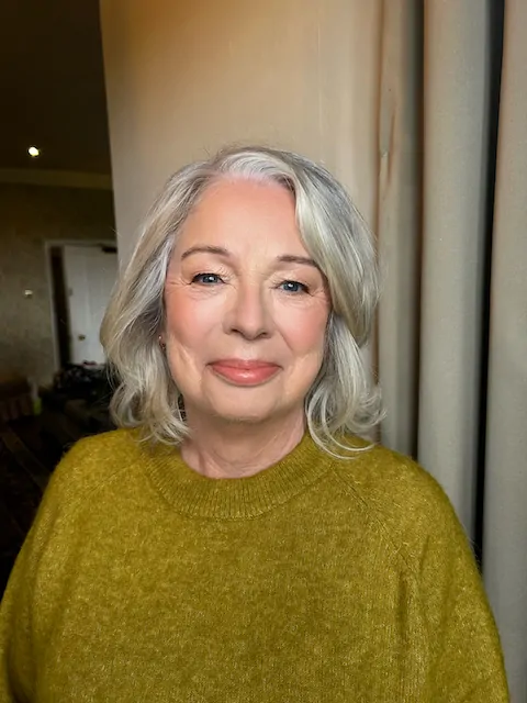 A mature bride with grey hair smiling gently indoors, showcasing natural wedding makeup and soft lighting.