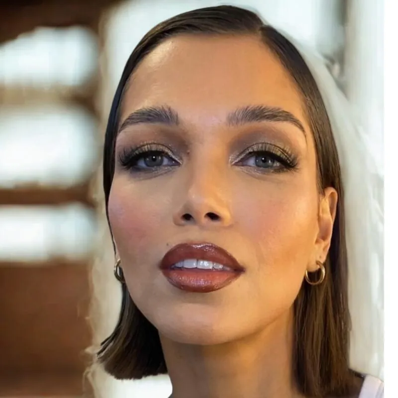 A close-up portrait of a bride displaying polished dewy makeup, glossy dark lips, fluttering lashes, and sleek short hair.
