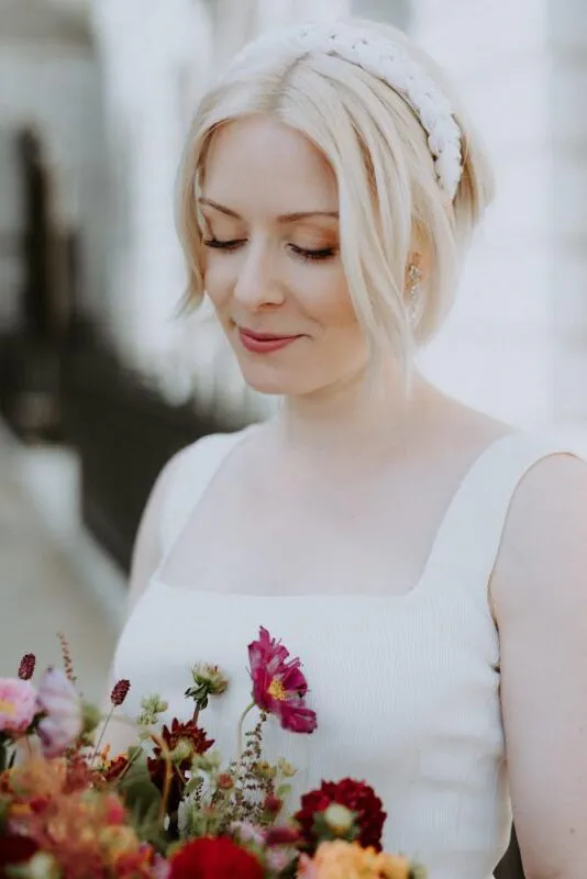 A blonde bride wearing a square neckline dress and a braided headband gazes down tenderly at a colourful wildflower bouquet.